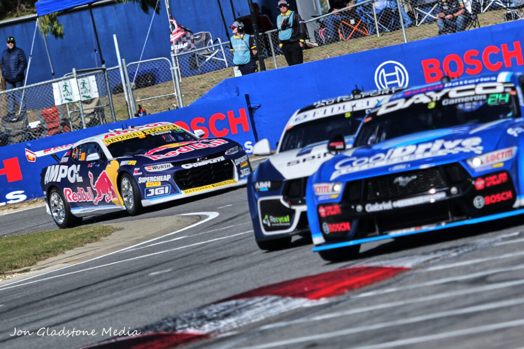 Three racecars in the photo, two close and out of focus with the third in focus and in the far-left corner. Some people are in the background, watching the cars go past.